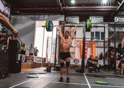 A muscular athlete performs a powerful overhead lift during a CrossFit competition indoors, showcasing strength and determination.